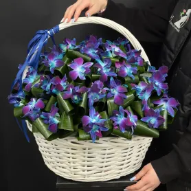 синие и фиолетовые орхидеи с зеленью в белой плетёной корзине